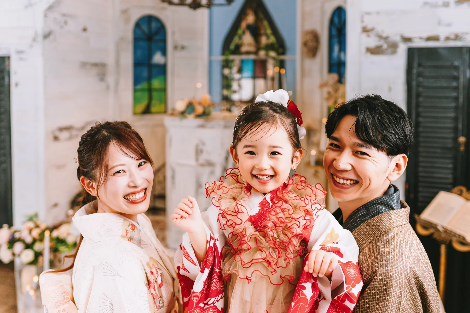 フリル衿と白い花の髪飾りで仕上げた赤と白の着物の3歳の女の子と、着物のお父さん・お母さん