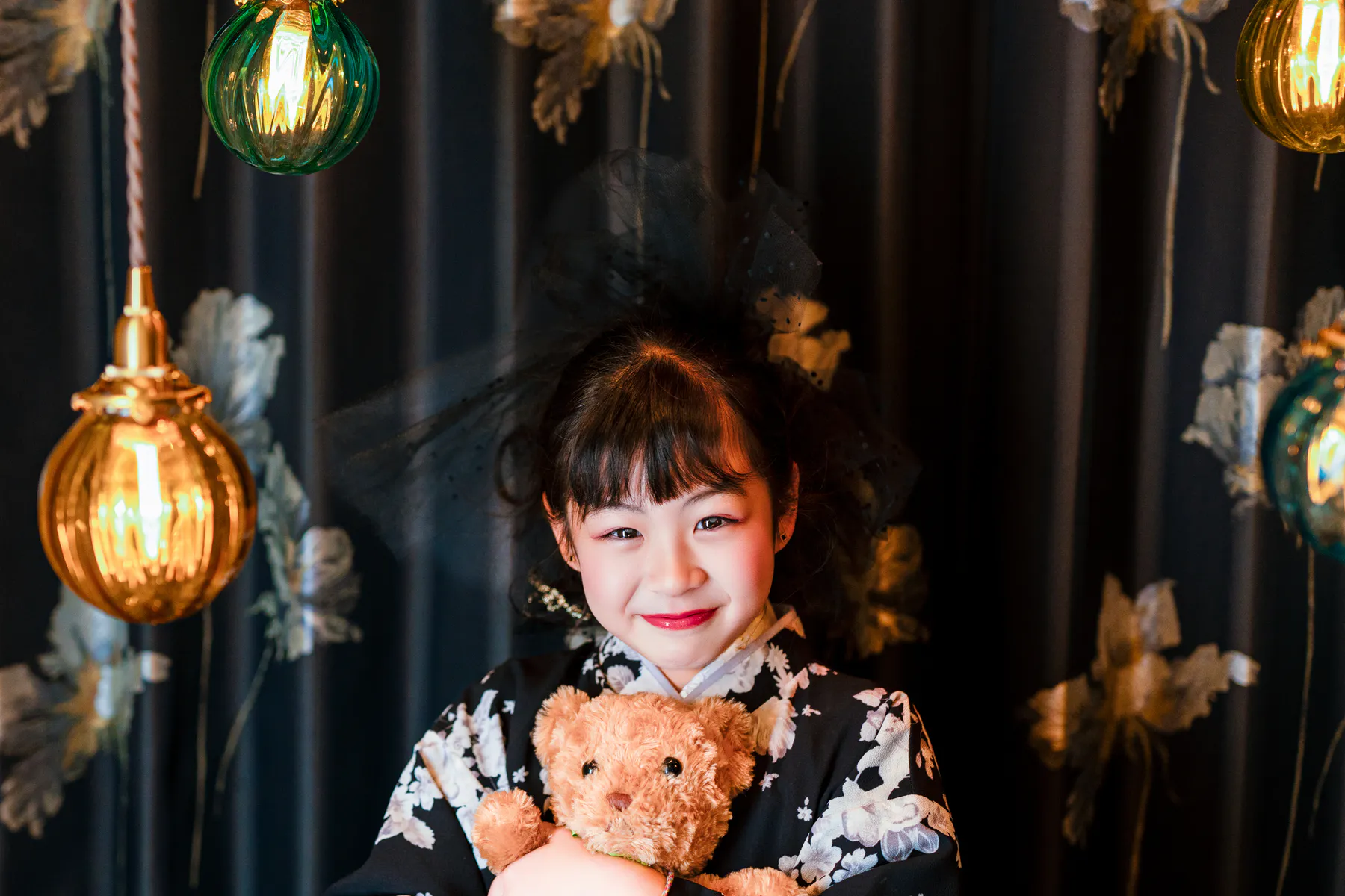 色硝子の灯りに囲まれ、くまを抱く黒い着物の子ども