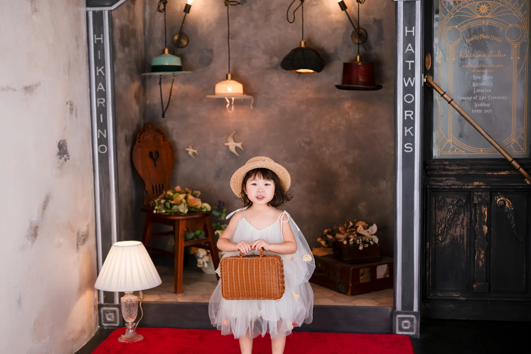 帽子屋の赤い絨毯に立つ、白いドレスとかごバッグの女の子