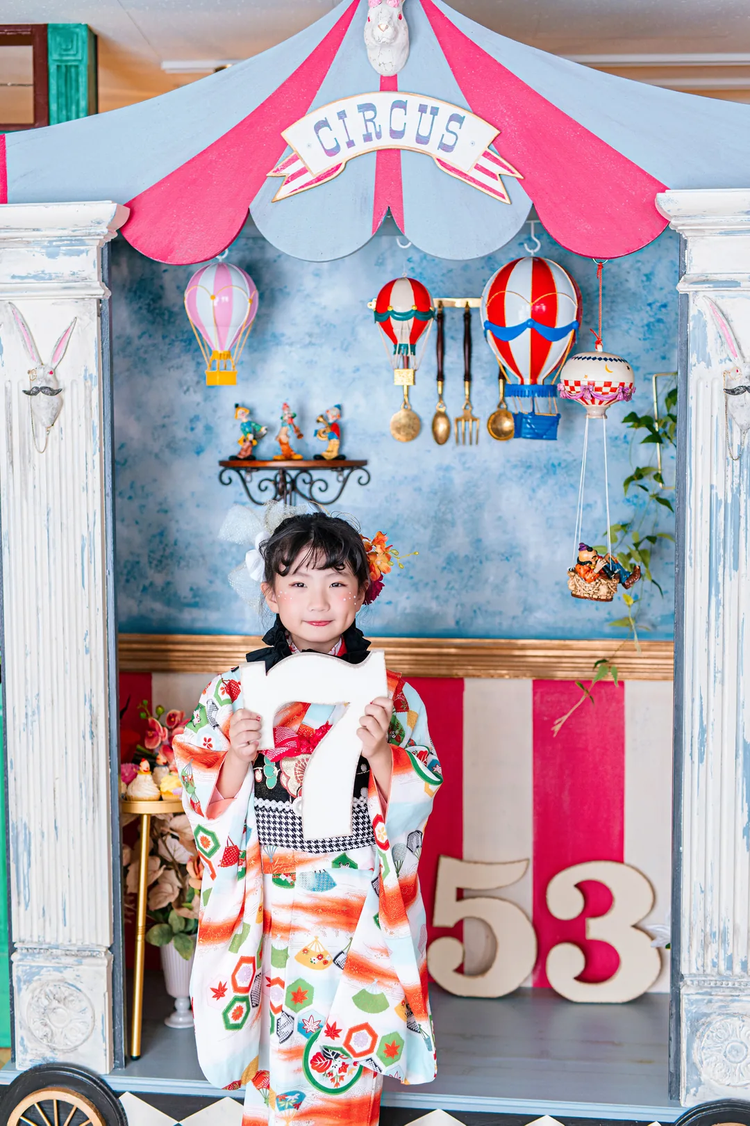 サーカスの小屋の前で数字の7を持つ、白地に扇の着物の女の子